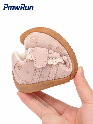 A baby's first shoe in light pink suede with teddy bear prints, lace detail, and a brown sole, held in a hand against a white background