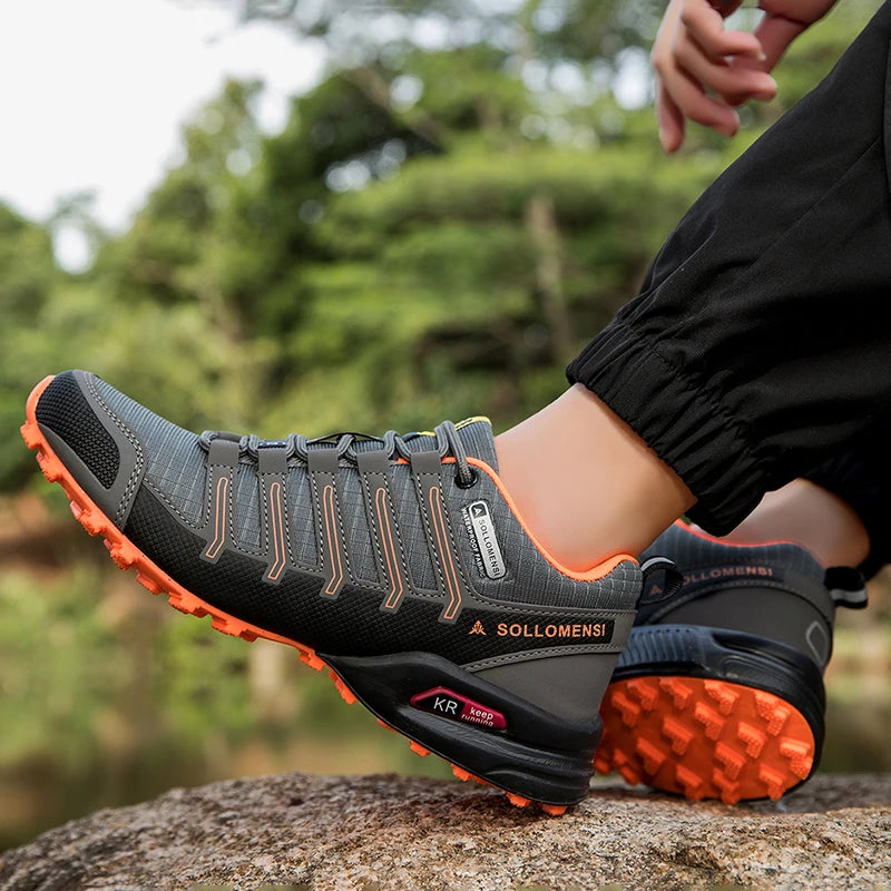 Person wearing gray and black hiking shoes with orange soles on a rock, blurred green background