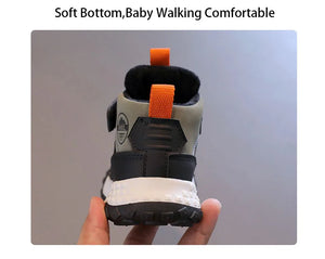 A baby bootie shown from the back, held by a hand against a plain gray background. The bootie has a light gray upper, black heel and sole, a white midsole, and bright orange pull tabs.