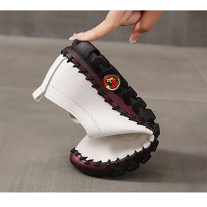 White lace-up split leather casual flats with round toes and rubber outsoles with black and red accents held by a hand on a gray background