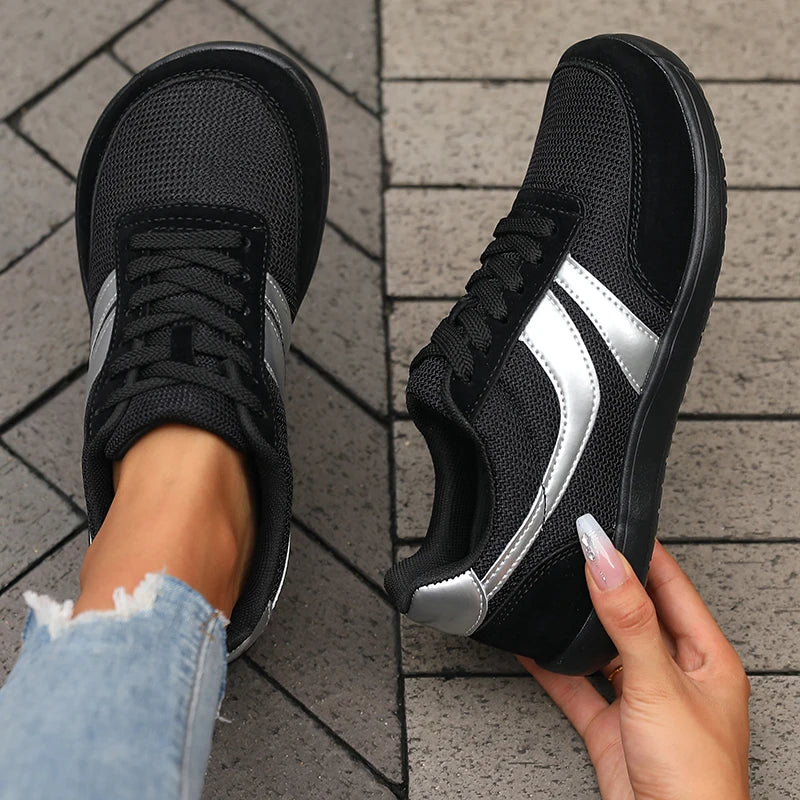 A pair of black athletic sneakers with metallic silver side stripes, shown with one shoe being worn on a foot and the other held by a hand over a gray tiled floor.