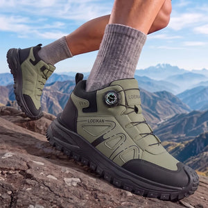 A pair of Loekan hiking boots in olive green and black, worn on a person's feet with gray socks while standing on a rocky mountain peak with a scenic mountain range in the background.
