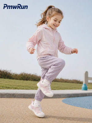 A young girl wearing a light pink windbreaker jacket, lavender joggers, and white running shoes jumps outdoors near a pool. The PmwRun brand logo is visible in the top left corner.