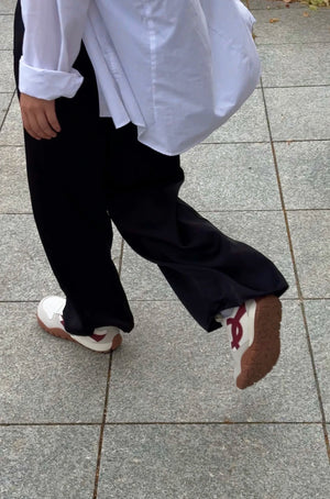 A person walking on a gray tiled outdoor pavement, wearing a white long-sleeve button-up shirt, loose black pants, and white sneakers with red accents.