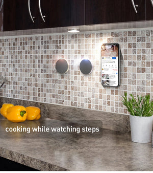 Smart kitchen setup with a phone displaying a recipe on a tiled wall above a countertop with peppers and a plant.