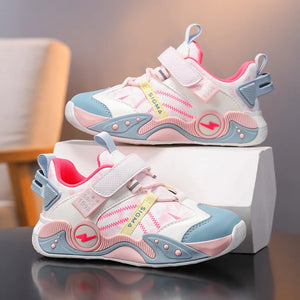 A pair of children's sneakers in white, pink, and light blue, displayed on a white box on a reflective dark surface with a blurred indoor background.