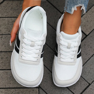 A pair of white low-top sneakers with black side stripes, shown being worn with a person's hand and legs visible against a gray tiled floor.