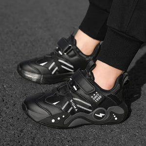 Black children's sneakers with white graphic accents, shown on a child's feet wearing black pants on an asphalt surface.