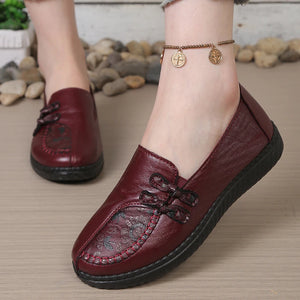 A close-up of a person wearing a burgundy leather slip-on loafer with decorative stitching and a lace-up front, shown outdoors on a stone path. A second matching shoe is visible in the background.