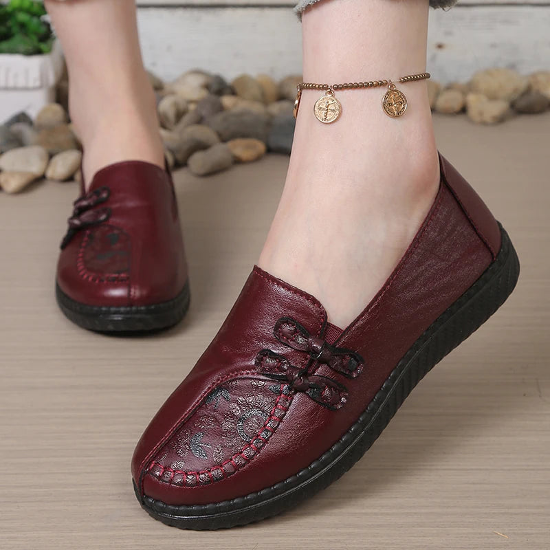 A close-up of a person wearing a burgundy leather slip-on loafer with decorative stitching and a lace-up front, shown outdoors on a stone path. A second matching shoe is visible in the background.