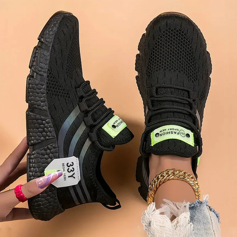 A pair of black knit athletic sneakers with gray side stripes and bright neon green accents, shown on a peach background. One shoe is held by a hand with a price tag attached, and the other is shown from the side.