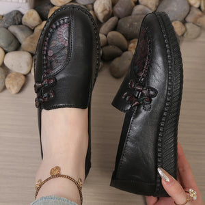 Black leather shoes with floral embroidery held by a person against a stone background