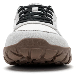 A close-up front view of a white and gray sneaker with black laces and a brown textured sole, shown on a white background.