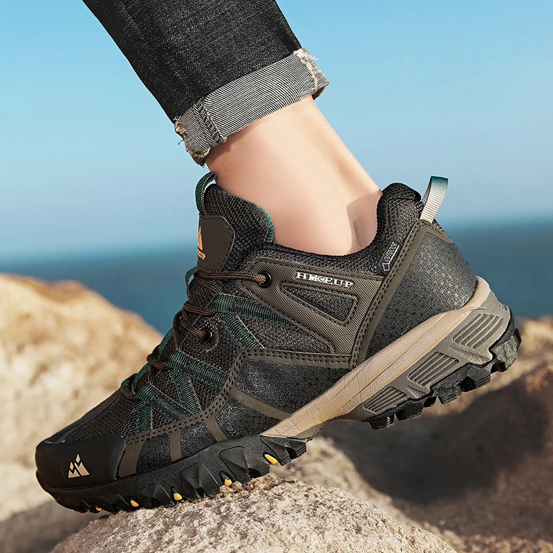A close-up of a person's foot wearing a dark gray and olive green technical hiking shoe with a rugged black sole, standing on a rocky outcrop with a blue sky and sea in the background.