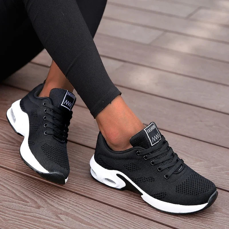 A woman modeling a black long-sleeve athletic top and matching black leggings with black sneakers, photographed against a clean white studio background.