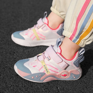 A close-up of a child wearing light pink, white, and light blue sneakers with hook-and-loop straps, standing on a dark asphalt surface. The child's pants have vertical stripes in cream, pink, blue, and yellow.