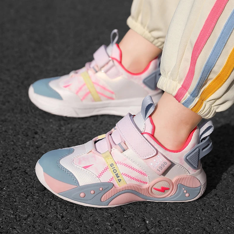 A close-up of a child wearing light pink, white, and light blue sneakers with hook-and-loop straps, standing on a dark asphalt surface. The child's pants have vertical stripes in cream, pink, blue, and yellow.