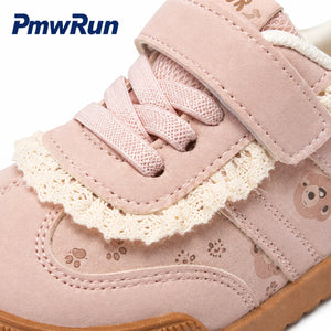 A close-up of a children's shoe in pale pink suede with cream lace trim, hook-and-loop straps, and small teddy bear face prints on the side.