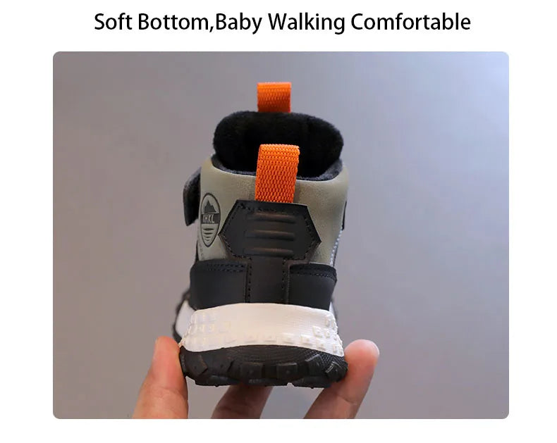A baby bootie shown from the back, held by a hand against a plain gray background. The bootie has a light gray upper, black heel and sole, a white midsole, and bright orange pull tabs.