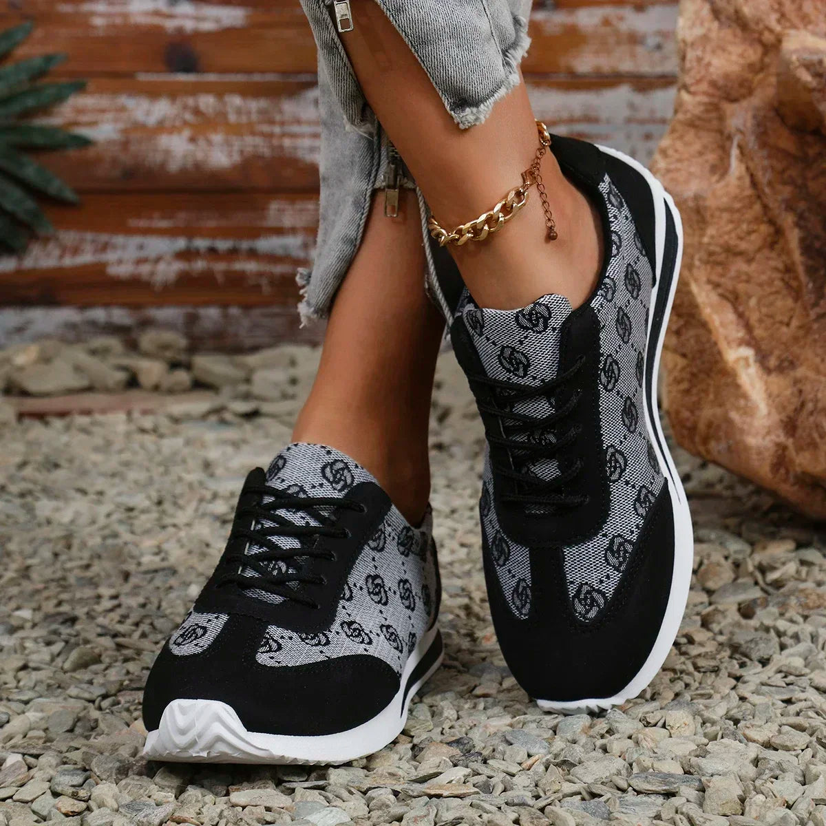 A person wearing black and gray patterned low-top sneakers with white soles, standing on gravel outdoors near a wooden wall and rock.