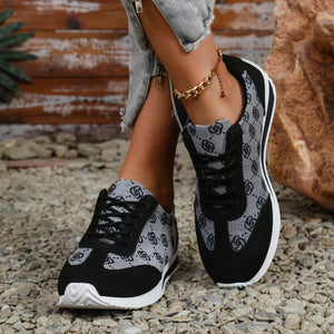 A person wearing black and gray patterned low-top sneakers with white soles, standing on gravel outdoors near a wooden wall and rock.
