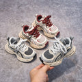 Four pairs of children's sneakers on a gray surface with a hand holding one pair.