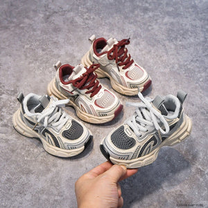 Four pairs of children's sneakers on a gray surface with a hand holding one pair.