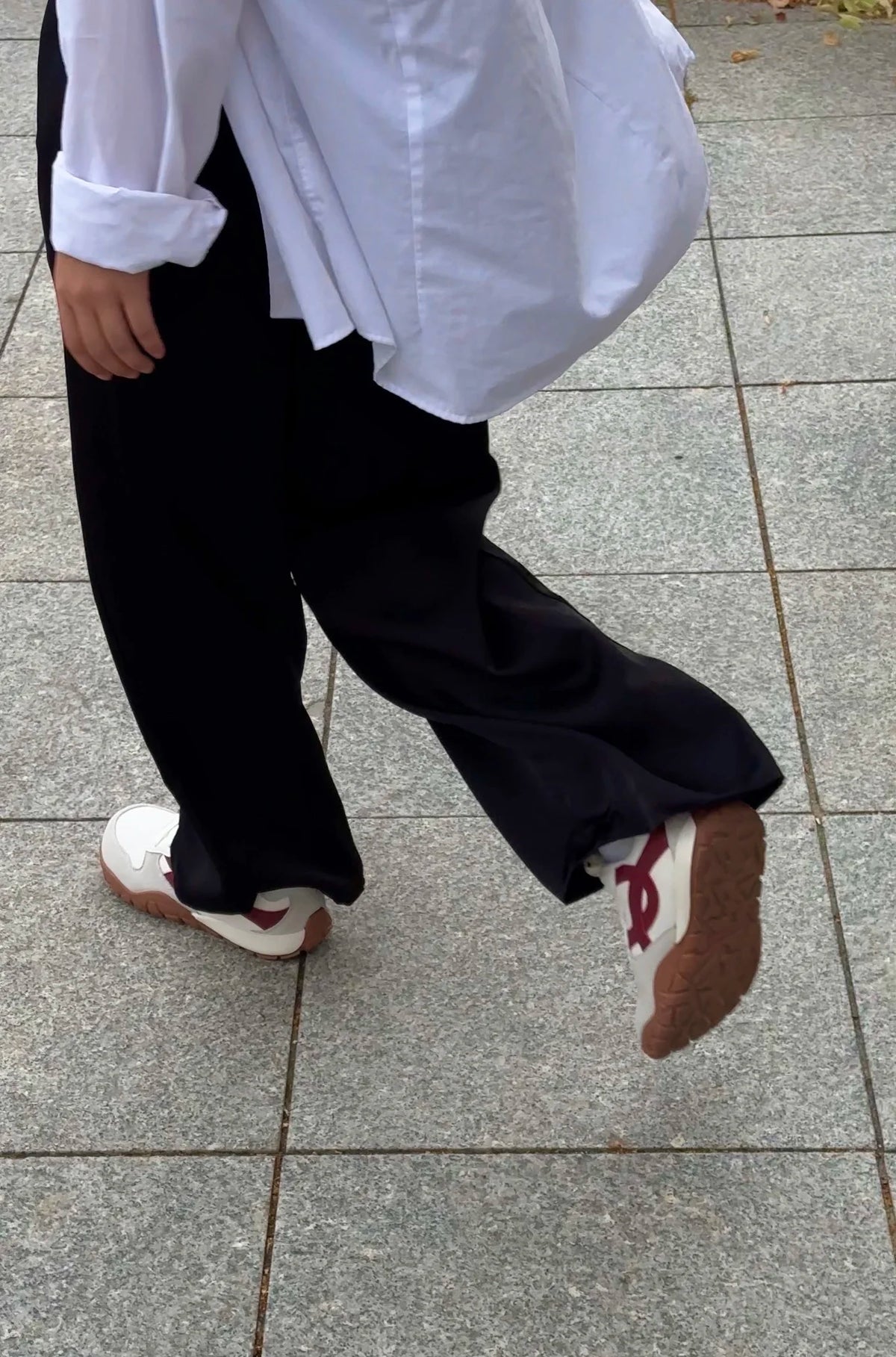 A person walking on a gray tiled outdoor pavement, wearing a white long-sleeve button-up shirt, loose black pants, and white sneakers with red accents.