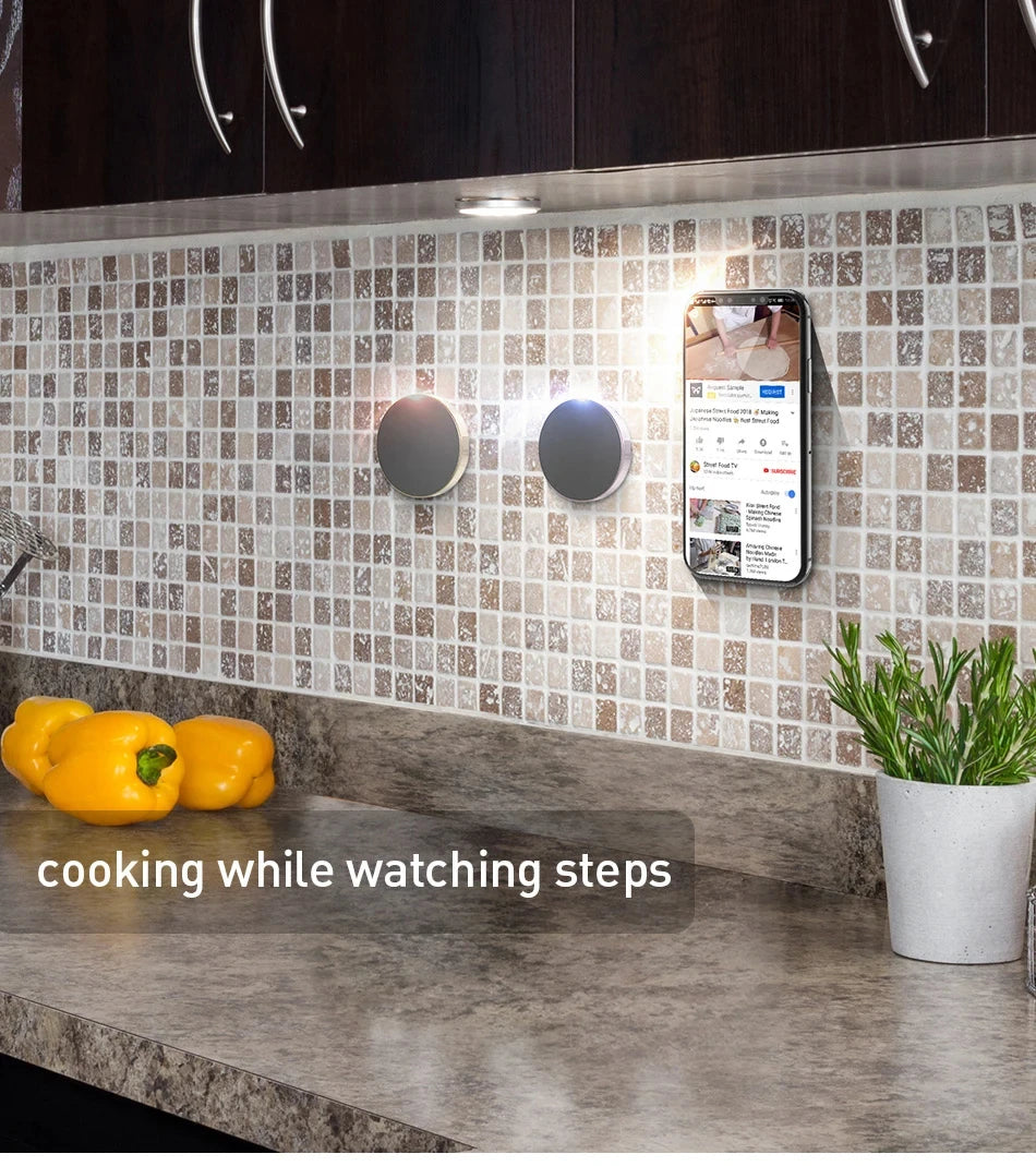 Smart kitchen setup with a phone displaying a recipe on a tiled wall above a countertop with peppers and a plant.