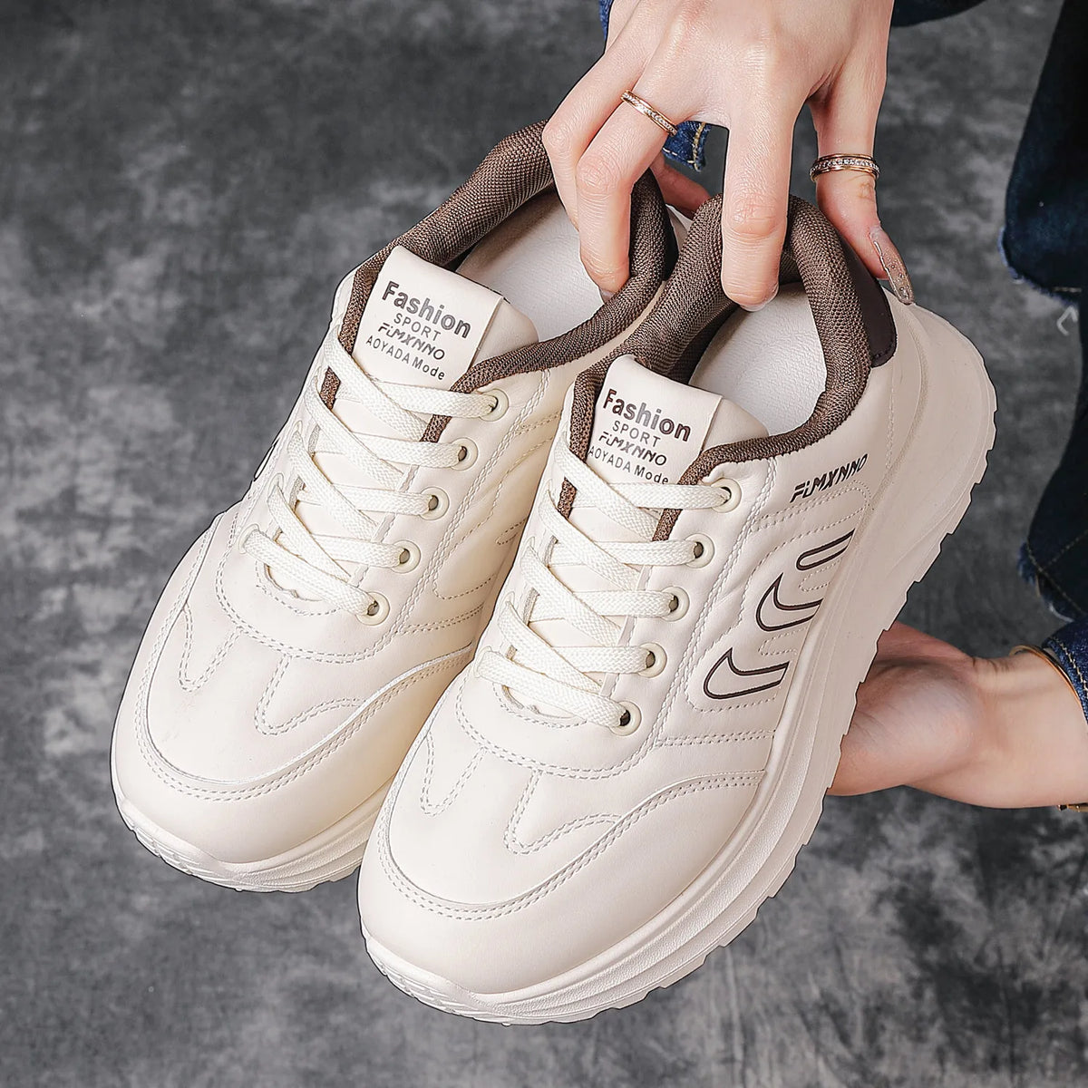A pair of chunky low-top sneakers in off-white and brown, held by a person's hands against a gray concrete floor. The shoes feature brown collar lining, brown laces, and black graphic accents on the side.