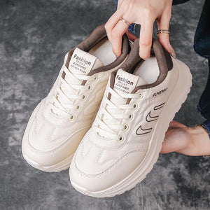A pair of chunky low-top sneakers in off-white and brown, held by a person's hands against a gray concrete floor. The shoes feature brown collar lining, brown laces, and black graphic accents on the side.