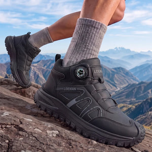 A person wearing black Loeikan hiking boots with gray crew socks, standing on a rocky mountain path with a scenic mountain valley and blue sky in the background.