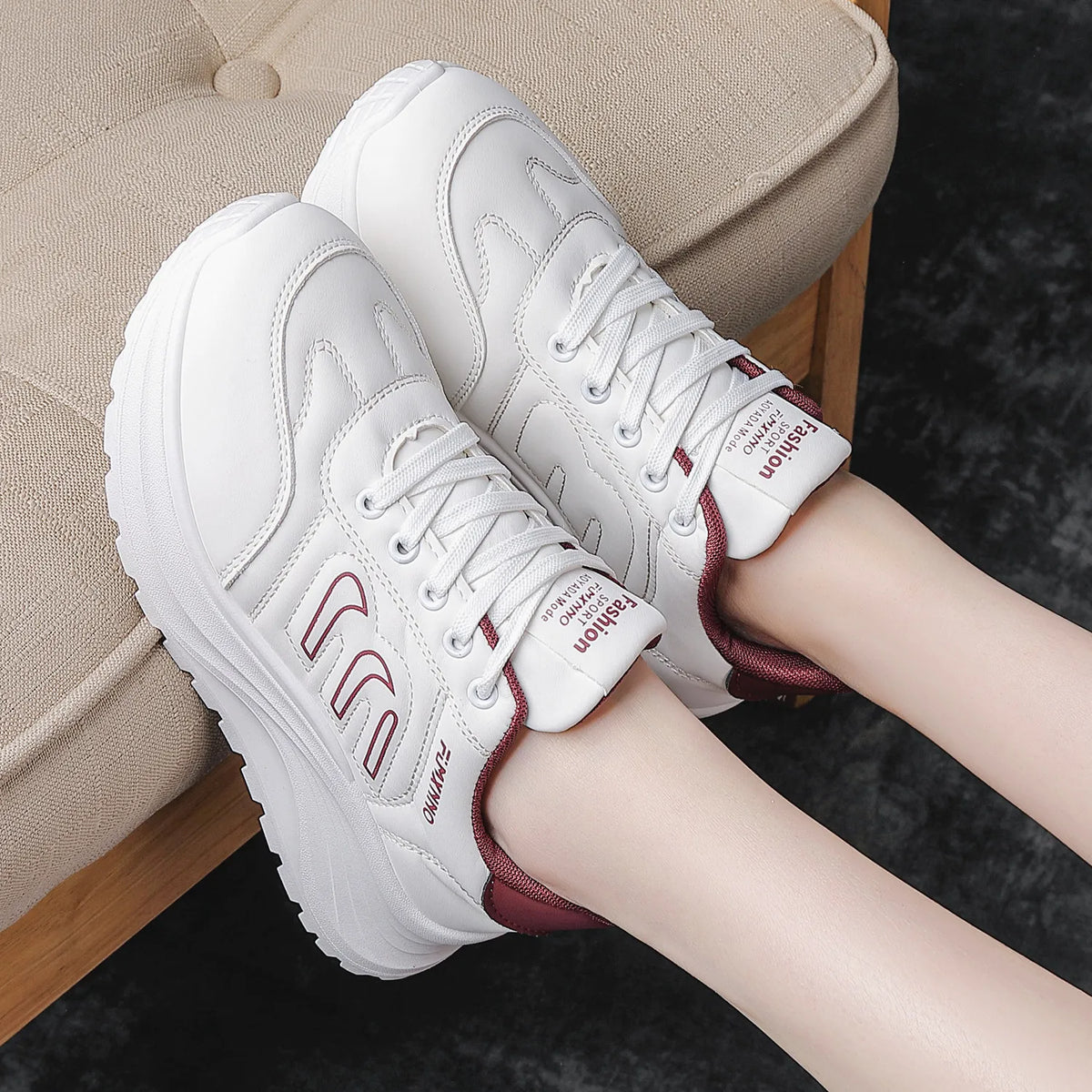 White platform sneakers with burgundy accents, shown on a model's feet sitting on a beige upholstered bench.