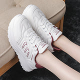 White platform sneakers with burgundy accents, shown on a model's feet sitting on a beige upholstered bench.