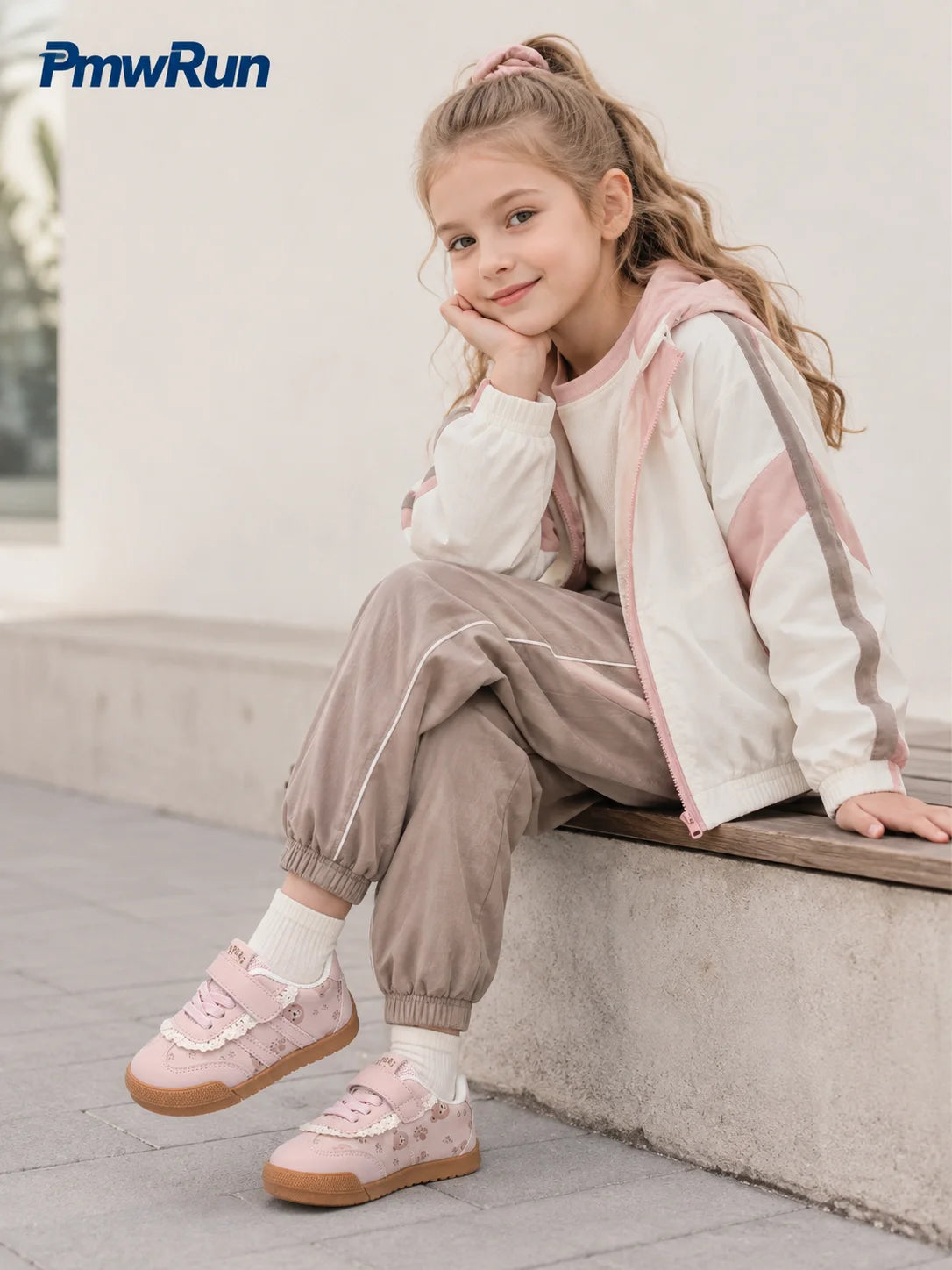 A young girl sits on a concrete ledge wearing a white and pink zip-up hoodie, matching jogger pants, and pink sneakers