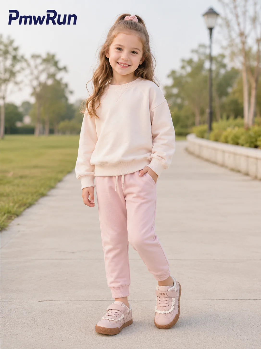 A young girl stands on a paved path in a park wearing a light pink sweatshirt and matching jogger pants, with pink sneakers