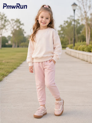 A young girl stands on a paved path in a park wearing a light pink sweatshirt and matching jogger pants, with pink sneakers