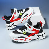 A pair of white, black, and red low-top sneakers with mesh panels, hook-and-loop straps, and wavy sole details, displayed on a clean studio background with a white geometric block.