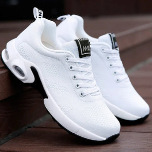 A pair of white athletic sneakers with black accents, photographed outdoors against a dark wooden surface. The shoes feature a breathable mesh upper, white laces, a chunky white sole with visible air pockets, and a black heel accent.