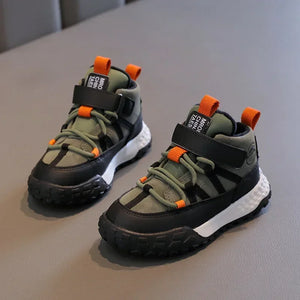 A pair of children's mid-top sneakers in olive green and black with bright orange accents, displayed on a gray textured surface.