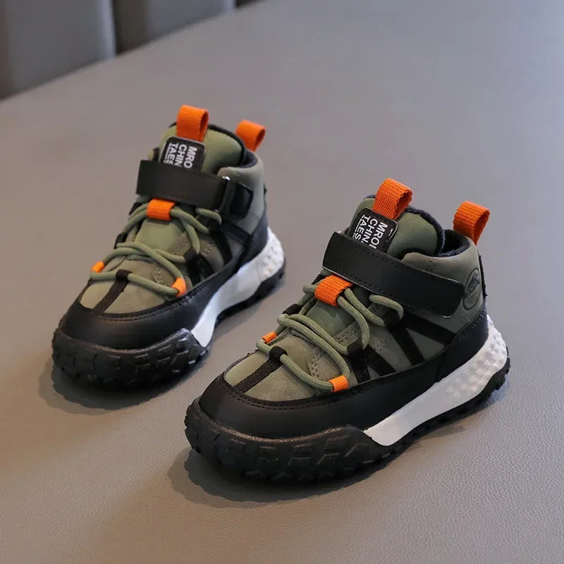 A pair of children's mid-top sneakers in olive green and black with bright orange accents, displayed on a gray textured surface.