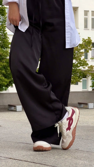 A person outdoors wearing loose black wide-leg pants, a white shirt, and chunky white sneakers with a burgundy side logo.
