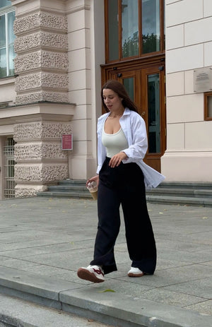 A woman walking outdoors in front of a stone building, wearing a white open-front shirt over a white tank top, loose black wide-leg pants, and white sneakers.