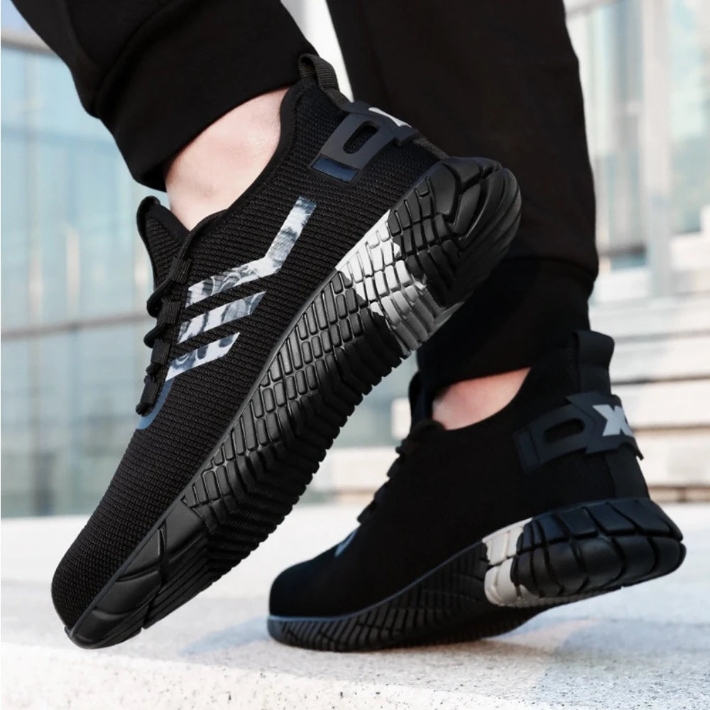 A pair of black work shoes with textured soles and reflective silver side stripes, shown being worn with black pants on a light concrete surface.