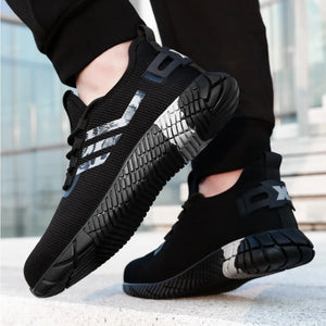 A pair of black work shoes with textured soles and reflective silver side stripes, shown being worn with black pants on a light concrete surface.