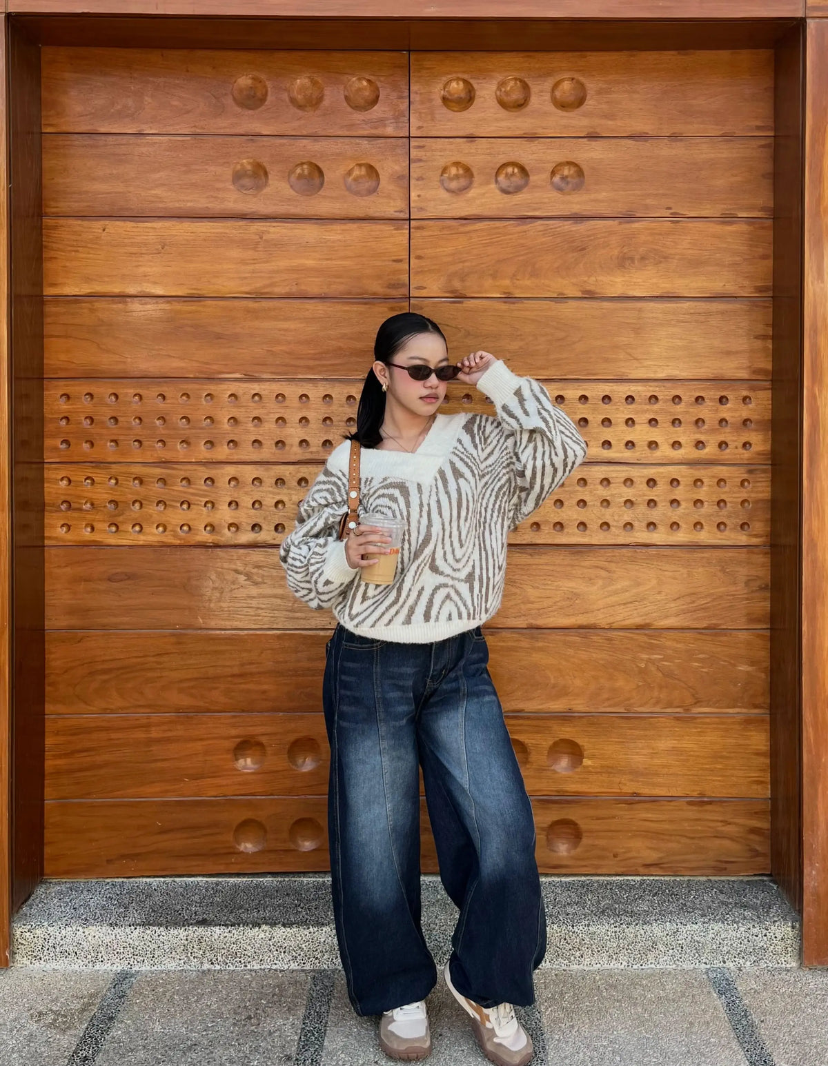 A woman stands in front of a large wooden door wearing a cream and black zebra-print knit sweater, dark wide-leg jeans, and beige sneakers, holding a small tan shoulder bag.