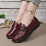Brown leather shoes worn by a person on a wooden floor with pebbles and plants in the background.