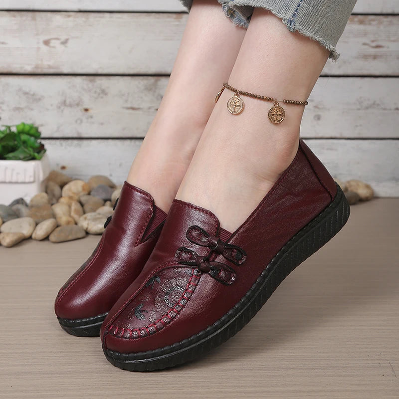 Brown leather shoes worn by a person on a wooden floor with pebbles and plants in the background.