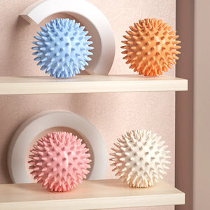 Four textured massage balls in blue, orange, pink, and white on a shelf against a beige wall.
