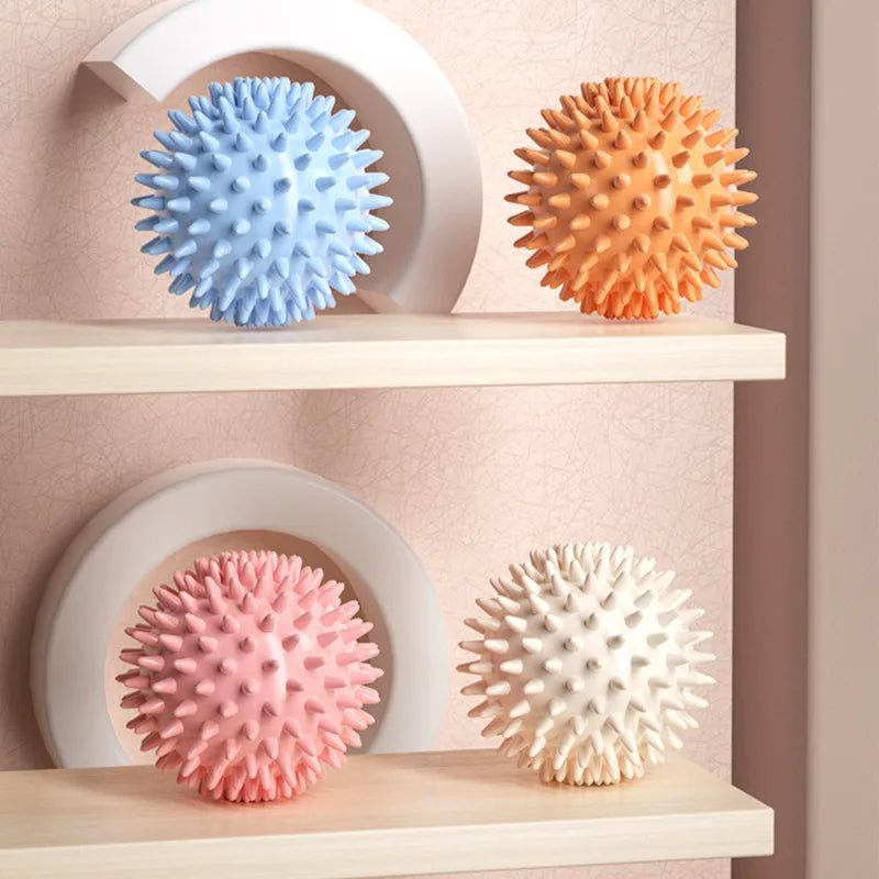 Four textured massage balls in blue, orange, pink, and white on a shelf against a beige wall.